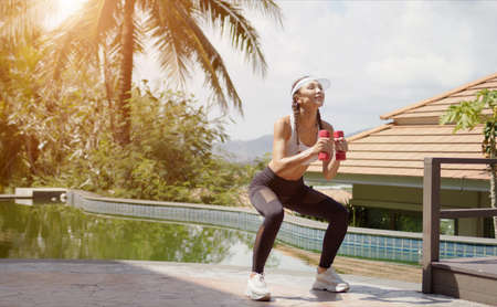 Full length fit lady in sportswear performing squats with dumbbells near swimming pool during fitness workout on sunny day on resortの写真素材