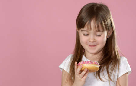 Studio portrait of a beautiful little girl holding a donut and smelling tasty flavour. Isolated on pink backgroundの写真素材