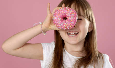 Young beautiful happy and excited blond girl 6 or 7 years old holding donut on her eyes looking through. Playing cheerful with cute smile while posing over ping background. Isolatedの写真素材