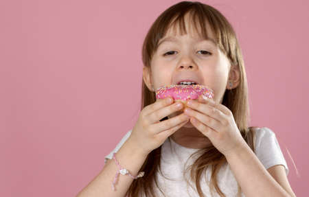 Cute young caucasian girl caught eating a sweet pink doughnut. Wearing white tshirt while standing isolated on pink background in studioの写真素材