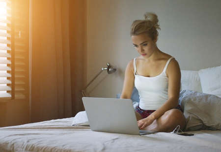 Pretty young woman in shorts and tank top sitting on comfortable bed and using laptop at sunsetの写真素材