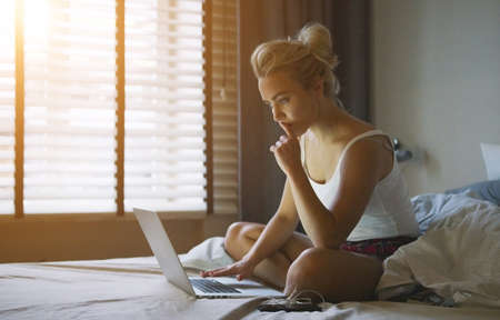 Beautiful young woman in pajamas sitting on bed with laptop and holding finger near lips while thinking.の写真素材