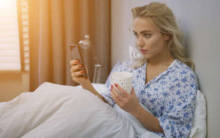 Beautiful young woman browsing smartphone and holding mug of hot beverage sitting on bed under warm blanket.の写真素材