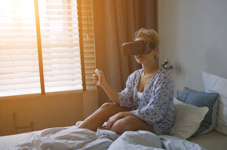 Young woman in shirt sitting on comfortable bed and using VR headset with controller at sunsetの写真素材