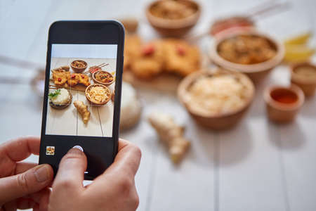 Woman taking a photo with smartphone of deep fried Crispy chicken in breadcrumbs served on white wooden table with salad, spices, ingredients and noodles. Asian food concept.の写真素材