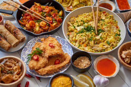Asian food served. Plates, pans and bowls full of tasty oriental dishes. Noodles chicken stir fry and vegetables ingredients with spices, sauces and chopsticks on whie wooden background.の写真素材