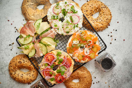 Delicious Bagel sandwiches with creamy cheese, ham, hummus, salmon and vegetables. Served on black grill on white rustic table. Healthy homemade food concept.の写真素材