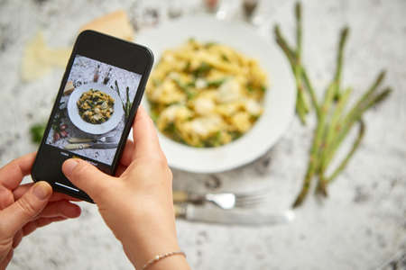 Woman making a photo with a smarpthone of homemade tagliatelle pasta with creamy ricotta cheese sauce and asparagus served white ceramic plate. Igredients on side. Top viewの写真素材