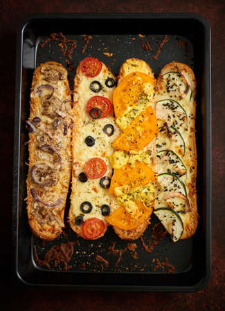Composition of hot tasty baked sandwiches with various toppings. Cheese, tuna, mozarella, olives, spices, zucchini, tomato. Delicious homemade food. Top view.の写真素材