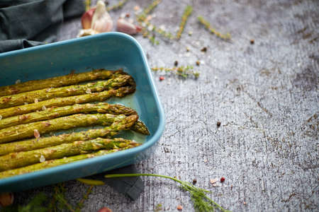 Roasted asparagus seasoned with salt, pepper, garlic and decorated with fresh herbs. Served in blue heatproof dish. Placed on gray stone background.の写真素材