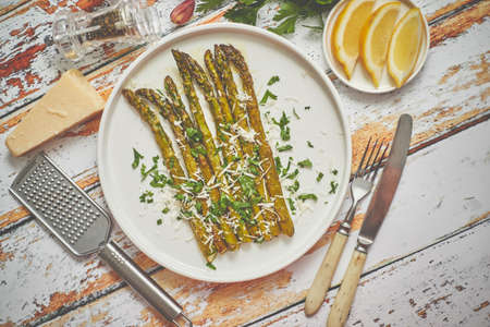 Delicious roasted asparagus served on white ceramic plate. With parmesan cheese, parsley and lemon. Placed on wooden table.の写真素材