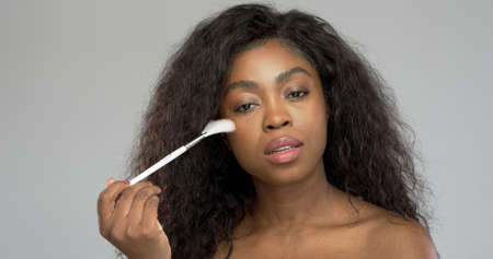 Pretty black female looking at camera and using brush to apply powder on cheek against gray backgroundの写真素材