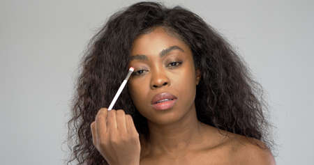 Beautiful black woman looking at camera and smearing eyeshadow on eyelid while applying makeup against gray backgroundの写真素材