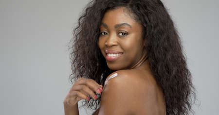 Side view of African American female with curly hair smiling and looking at camera while applying cream on shoulder against gray backgroundの写真素材
