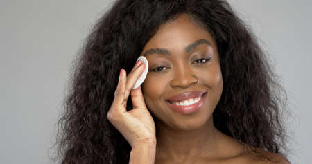 Cheerful black woman smiling and using cotton pad to clean face from makeup during skin care routine against gray backgroundの写真素材