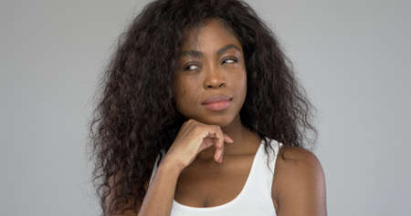 Beautiful African American woman with curly hair touching chin and looking away while pondering against gray backgroundの写真素材
