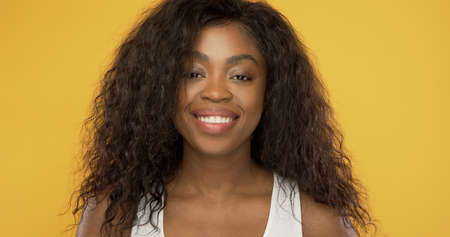 Beautiful African American lady with curly hair cheerfully smiling and looking at camera against bright yellow backgroundの写真素材