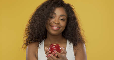 Happy black woman with curly hair smiling for camera and showing ripe red apple against bright yellow backgroundの写真素材