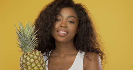 Happy black female with fresh pineapple friendly smiling and looking at camera against bright yellow backgroundの写真素材