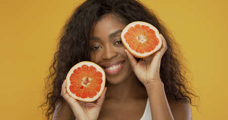 Optimistic African American woman with curly hair smiling for camera and demonstrating halves of ripe juicy grapefruit on summer day against yellow backgroundの写真素材