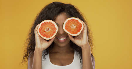 Happy black female with curly hair smiling and holding halves of fresh grapefruit near eyes while having fun against yellow backgroundの写真素材