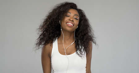 African American lady in white top smiling and closing eyes while listening to music in earphones against gray backgroundの写真素材