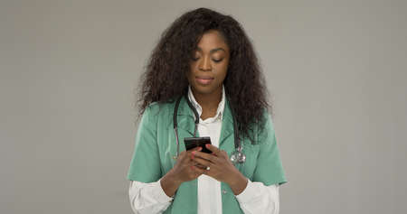 African American female doctor with stethoscope texting message on smartphone during break in work against gray backgroundの写真素材