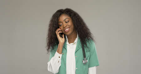 Cheerful African American woman in medical uniform smiling and looking away while having smartphone conversation against gray backgroundの写真素材