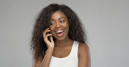 Excited ethnic female with curly hair smiling and looking away while answering phone call against gray backgroundの写真素材