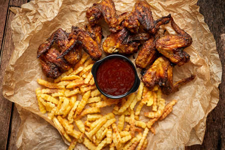 Appetizing roasted chicken wings and french fries with barbecue dip, served on baking paperの写真素材