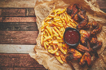 Appetizing roasted chicken wings and french fries with barbecue dip, served on baking paperの写真素材