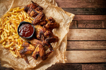 Appetizing roasted chicken wings and french fries with barbecue dip, served on baking paperの写真素材