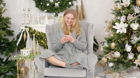 Smiling trendy woman in gray cozy sweater sittin on comfortable armchair and sending text messageの写真素材
