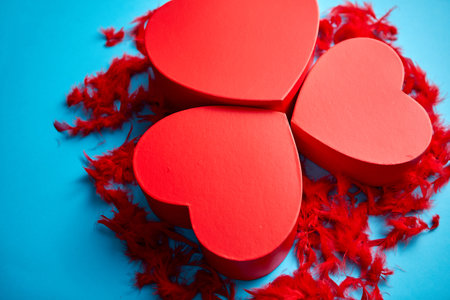 Three red, heart shaped gift boxes placed on blue background among red feathersの写真素材