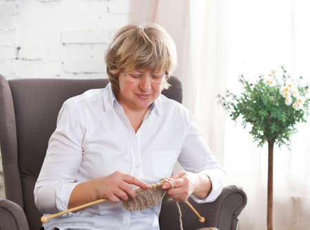 Portrait of a smiling middle age woman knitting on spokes at homeの写真素材