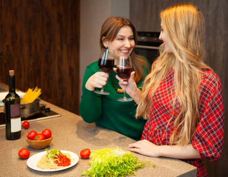 Two beautiful young women friends drinking red wine together in a kitchenの写真素材