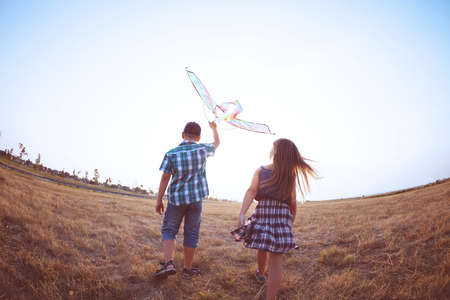 Happy boy and little girl running with bright kite on a meadow in a sunny eveningの写真素材
