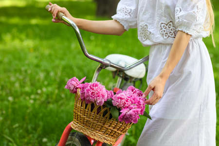 Beautiful blond woman wearing a nice dress in park with bicycle carrying a beautiful basket of peony flowers. Pretty blonde girl with retro look, bike and basket with flowers. Close upの写真素材