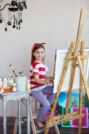 Little artist girl holding a paintbrush and looking over a canvas on an easel. Concept of creativity and funの写真素材