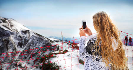 Blond woman photographing winter landscape mountains and snow with cell phoneの写真素材