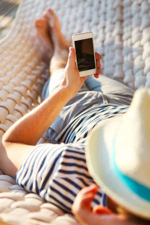 Lazy time. Man in hat in a hammock with mobile phone on a summer dayの写真素材