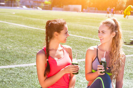 Two happy women drinking vegetable smoothie after fitness running workout on stadium. Fitness and healthy lifestyle conceptの写真素材
