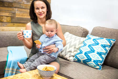Young happy mother drinking coffee and her child eating cookies at the living room at home