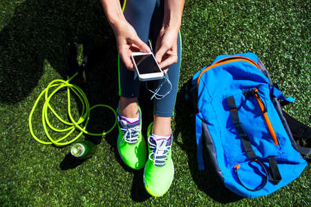 Sporty woman holding phone with empty screen sitting on the green grass with jump rope, backpack and drink bottleの写真素材