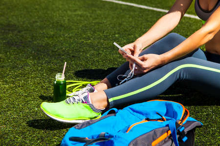 Sporty woman holding phone with empty screen sitting on the green grass with jump rope, backpack and drink bottleの写真素材