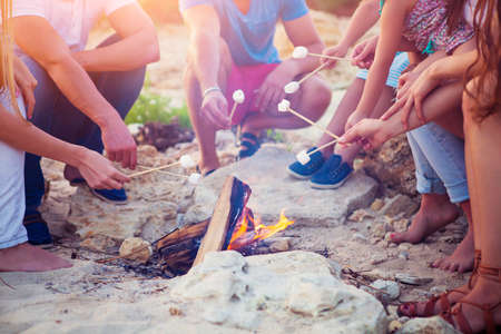 Friends sitting on the sand at the beach in circle with marshmallow on the beachの写真素材