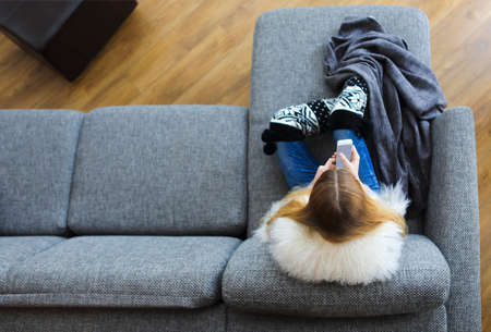 Woman lying on sofa while writing a text message on her cell phoneの写真素材