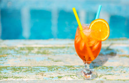 Glass of orange cocktail with slice of orange and straw near poolの写真素材