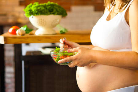 Young pregnant woman eating fresh vegetable salad at the kitchen. Pregnant nutritionの写真素材