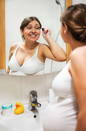 Young pregnant woman applying mascara and looking at mirror in bathroomの写真素材